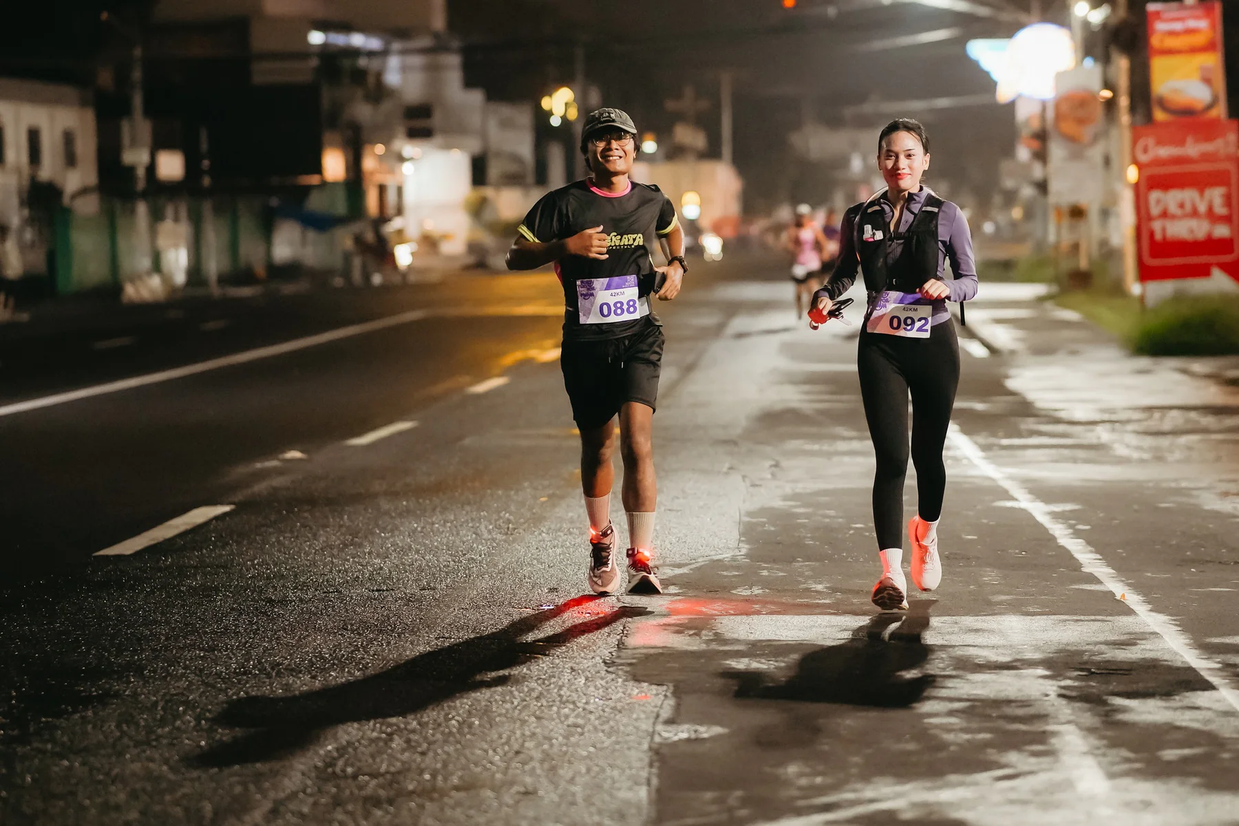 Two runners smiling as they move together along a wet road at night